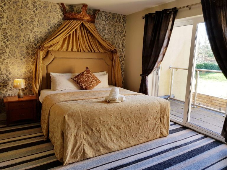 Beautifully decorated double room at Killarney Inn, County Kerry, Ireland, featuring an elegant gold bedspread, crown-style headboard with draped fabric, and large glass doors opening to a private balcony with garden views. The room includes warm lighting, rich damask wallpaper, striped carpet, and a cozy side table — offering a royal touch in a comfortable, affordable stay near Killarney town centre and Killarney National Park.