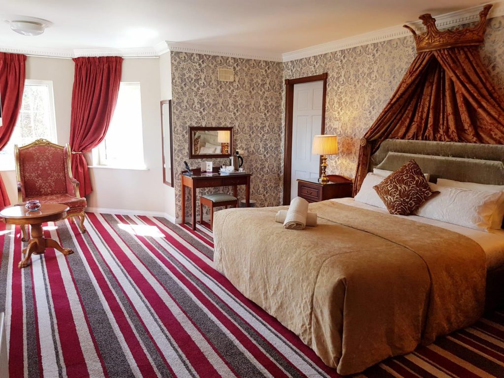 Luxurious and spacious room at Killarney Inn with a king-size bed, vintage-style red armchair and coffee table, striped red carpet, and opulent crown-draped headboard. Includes a writing desk, tea/coffee station, and large windows offering ample natural light. Perfect for guests seeking comfort and elegance in the heart of Killarney.