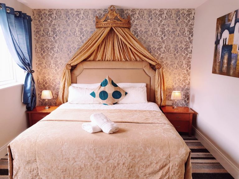 Stylish double room at Killarney Inn with regal gold bedding, blue accent curtains, and a unique crown-inspired canopy above the headboard. A centrally placed polka-dot decorative pillow adds a modern flair. Perfect for couples or solo travellers exploring Killarney, the room includes bedside lighting, patterned wallpaper, and a welcoming, well-lit atmosphere close to the Ring of Kerry and local attractions.