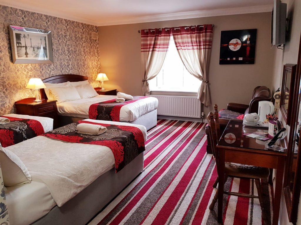 Spacious family or group room at Killarney Inn, featuring one double bed and two single beds with coordinated red and black bedding, set against ornate wallpaper. The room includes a work desk, flat-screen TV, tea/coffee amenities, and large windows with striped curtains. Ideal for families or small groups visiting County Kerry and enjoying the comfort of a stylish, centrally located guesthouse.