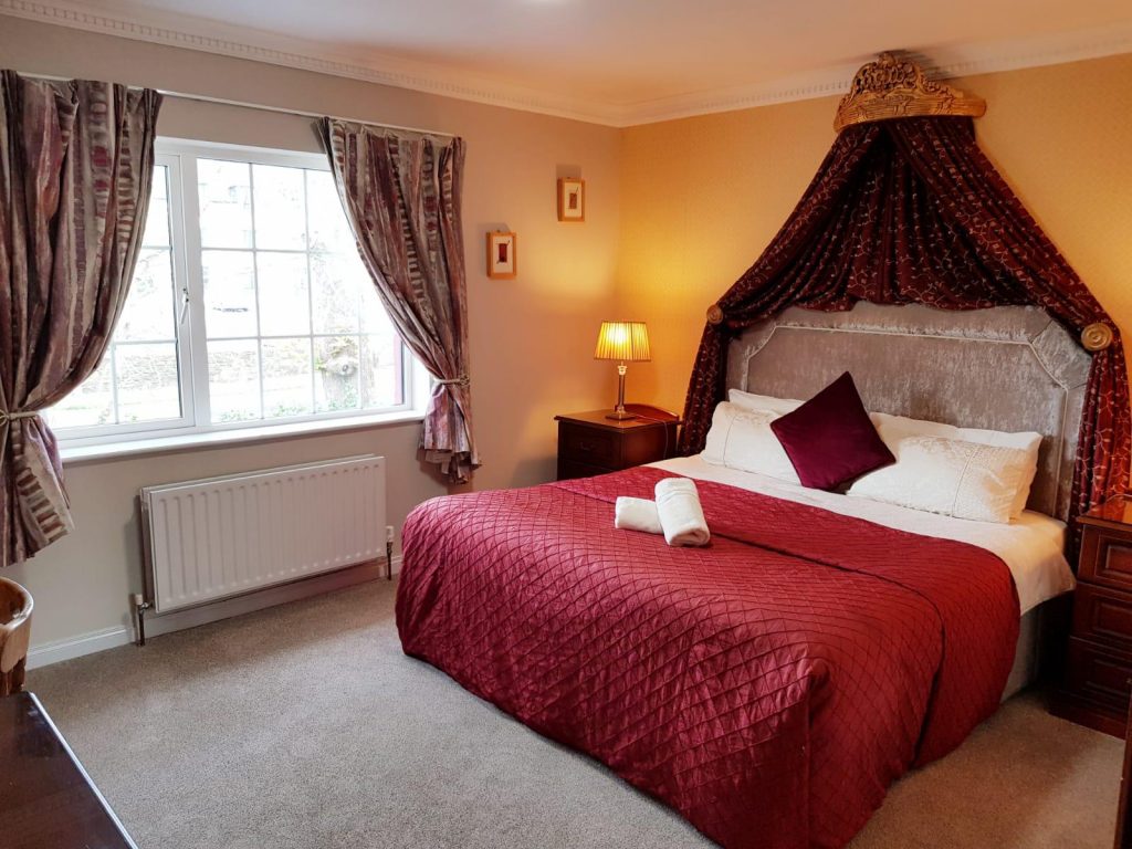 Elegant room at Killarney Inn with plush red bedding, deep burgundy canopy drapery above the bed, and traditional wooden furnishings. A cozy and romantic atmosphere perfect for couples seeking a warm and stylish retreat while exploring the scenic landscapes of Killarney National Park, Muckross House, and the Ring of Kerry.