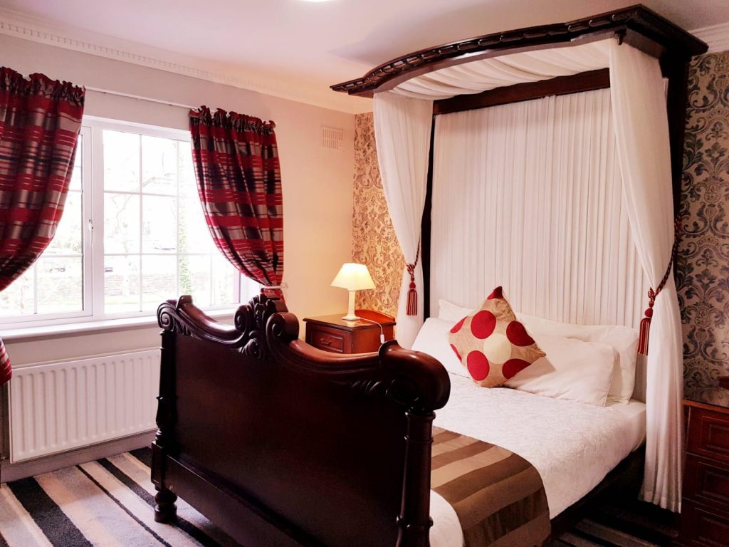 Charming room at Killarney Inn featuring a beautifully carved wooden canopy bed with white drapes and a red polka-dot cushion. The room includes large windows with red striped curtains, bedside tables with lamps, and elegant wallpaper. A cozy and unique setting for a comfortable stay near Killarney’s town centre and natural attractions.