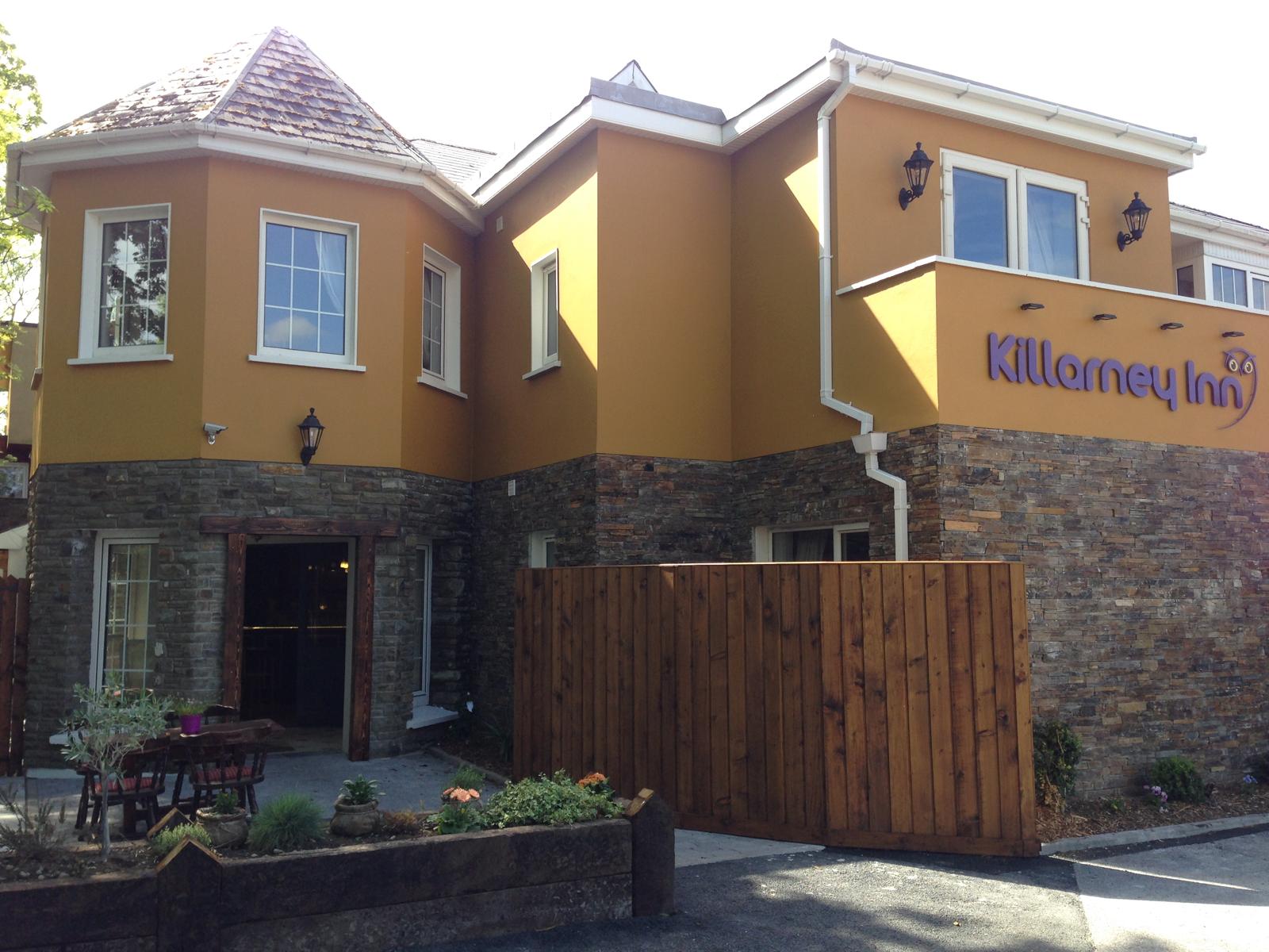 Front exterior of Killarney Inn, a welcoming and affordable guesthouse in Killarney, County Kerry. The building features warm yellow walls, natural stone accents, and a distinctive turreted corner with white-trimmed windows. The signage and well-maintained entrance highlight its easy-to-access location near Killarney National Park and the town centre.