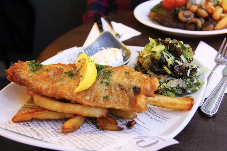 Delicious plate of traditional fish and chips served in a charming Irish restaurant in Killarney, County Kerry. The photo features golden, crispy battered fish, thick-cut seasoned chips, a side of mushy peas, and a wedge of lemon, beautifully plated with tartar sauce and a fresh garnish. Set against a cozy, rustic pub-style interior, the dish represents classic Irish comfort food at its best. A popular meal for tourists and locals alike, especially after a day exploring Killarney National Park, Muckross House, or the Ring of Kerry. Perfect for those seeking authentic Irish cuisine in a warm, welcoming setting.