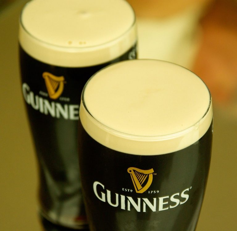Close-up view of two freshly poured pints of Guinness stout beer with creamy, thick heads, served in iconic branded glasses on a rustic wooden pub table. The rich dark color and smooth foam showcase the classic Irish beverage, known for its deep roasted flavor and velvety texture. Perfectly poured and ready to enjoy, this image evokes the warmth of traditional Irish hospitality, pub culture, and friendly gatherings. Often found in cozy pubs throughout Killarney and across Ireland, Guinness is a must-try for visitors seeking an authentic Irish drink experience.