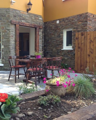 Charming outdoor seating area at Killarney Inn with wooden chairs and tables surrounded by flowers, greenery, and stone-clad walls. A peaceful space to enjoy coffee or unwind after a day exploring Killarney National Park, the Lakes of Killarney, or the Ring of Kerry. Ideal for guests seeking a mix of nature and comfort.